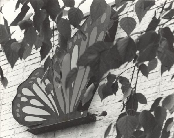 Flat sculpture or sign of a monarch butterfly attached to the side of a brick wall. Leaves are hanging from branches of a tree from the upper right and partially obscure the butterfly and wall.