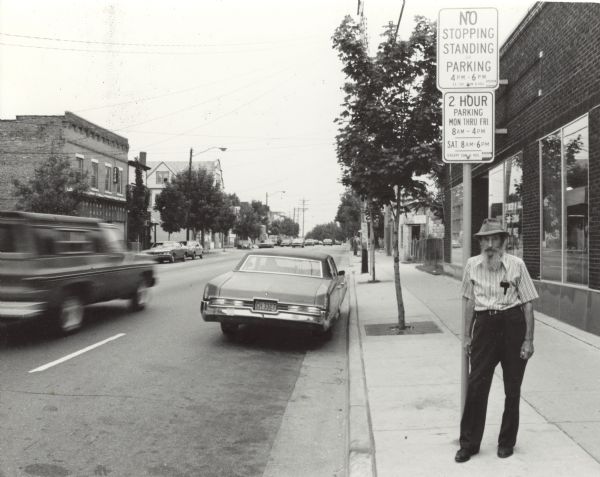 View down a curb towards a man standing on the sidewalk on the right leaning against a parking sign. He is wearing a hat and a striped shirt tucked into black pants. A truck is driving down the street and cars are parked along the curbs. Across the street on the left is the American Legion Post #501.