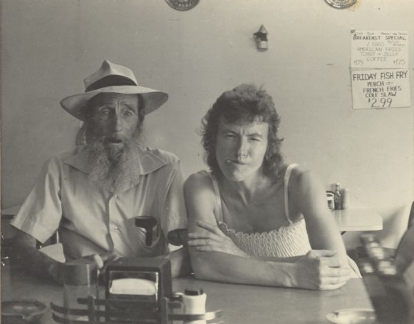 A man and a women sitting at the counter of a diner posing for the camera. The man has his eyes wide open and mouth slightly open. The woman is leaning cross-armed on the counter and squinting her eyes. She has her tongue sticking partially out of the right side of her mouth. Two paper signs are taped to the wall behind them listing the breakfast special and advertising the Friday fish fry.