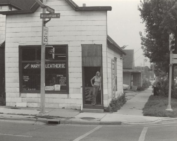 View across Williamson Street towards a woman standing in the open doorway of a shop with a sign that reads: "Crazy Mary's Leatherie." The building is on a corner with street signs mounted on a traffic signal that read: "Paterson St" and "Williamson St."