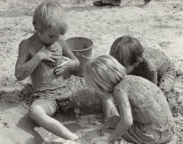 Three young boys playing in a small hole dug into the sand and filled with water. The boy's bodies and swimsuits are covered in muddy sand.