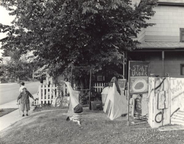 A woman is performing a headstand on her front lawn as an elderly woman is standing and watching from the sidewalk. A sign on the lawn reads: 'Start Under,' and a folded wall is painted with the words: 'TWA Dogs.' Other objects are placed in the lawn, including a small pyramid shaped structure. This was known as the Church of Anarchy, located at 1341 Williamson Street.