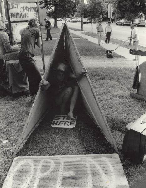 Elizabeth Was is crouching inside two triangular walls open on one side. In front of her are two painted signs lying on the grass and read: 'Step Here' and 'Open.' In the right foreground a TV is on the grass. Behind Was on the left is Miekal And standing near a sculpture of a Buddha, which is sitting on a cloth-covered stand and has a bird cage over its head. On the right near the sidewalk a window or a screen frames a flannel jacket. Next door a man is mowing the yard. Two men are walking up the sidewalk, and a woman is walking through the neighboring yard. This was known as the Church of Anarchy, located at 1341 Williamson Street.