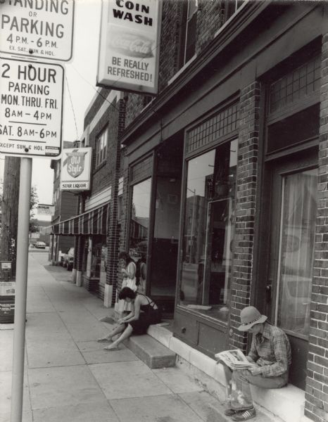 View down a sidewalk towards three people waiting outside of a coin laundromat on Williamson Street. Two women, one on steps of a doorway and another near a closed door, are sitting and reading newspapers. Another woman is standing and leaning against a window. Next door to the laundromat is Star Liquor.