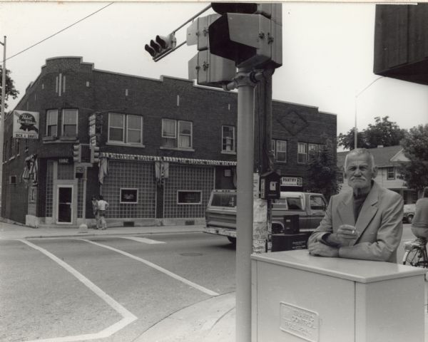 View from street of a man standing on the corner of Baldwin and Williamson streets resting his arms on a traffic control box. He is wearing a suit jacket and is holding a cigarette in his right hand. Across the street is the Crystal Corner Bar.