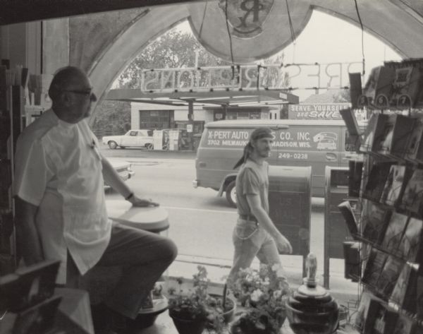View of Williamson Street from inside Shafer Pharmacy. The owner, Hugh Hessman, is standing by a large arched window looking out onto the street. He wears glasses and a white shirt. Flower pots are sitting on the ledge, and a rack of books is on the right. A neon sign in the window reads: "Prescriptions" (backwards) with the Rx symbol decorating the top of the window. Outside a man in jeans, t-shirt, and a headband is walking by on the sidewalk. Behind him are two mailboxes. A van is driving by on the street, with an advertisement for X-Pert Auto (part of the business name is obscured by the man). A gas station is across the street.