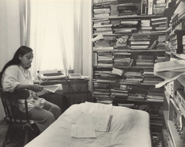 Angela Lobo-Cobb is sitting sideways in a chair in front of a desk at a window. She is looking down at papers in her hands. On the desk is a typewriter. Two bookshelves, filled with a radio, binders, boxes, and books on various subjects (mostly poetry, but there are a few cook books, history books, books on Eastern cultures/history and a few other subjects) stand against two walls in the room. In the foreground papers lay on a bed. Angela Lobo-Cobb has written and compiled a books of poetry, mostly on the experiences of minorities in the Midwest or Wisconsin.