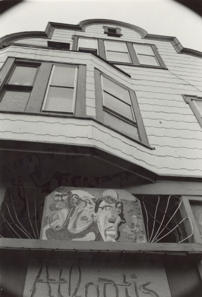 View looking up at the facade of a house. A sign at the bottom reads: "Atlantis" fashioned in wire. Above the sign is a painting that includes two male faces and a nude woman.