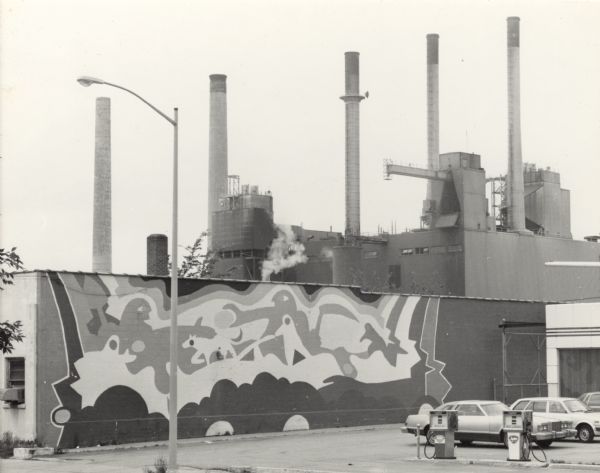 View across the street towards a mural on the side of an one-story brick commercial building. There are cars parked in the lot of a service station on the right. The industrial building is the Bill Doran Company, a floral distribution company. In the far background are smokestacks of the Madison Gas & Electric Company.