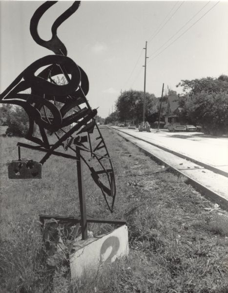 A metal abstract sculpture standing in the grass beside railroad tracks. The railroad tracks, and a road on the right, are leading into the background. On the other side of the road are houses, trees, and cars.