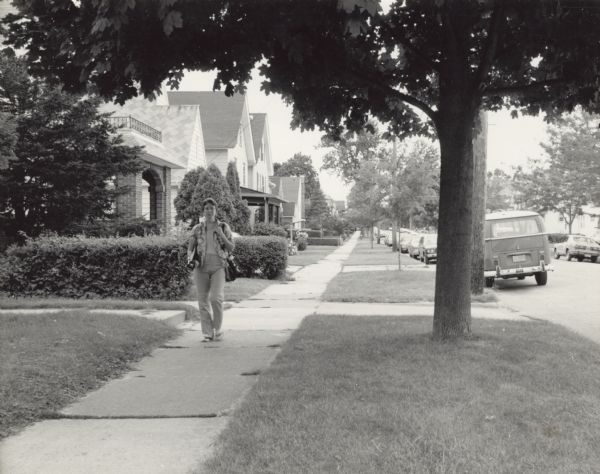 A woman, Lori Bose, is walking down a residential sidewalk with a tripod under her right arm. Cars are parked along the curb.