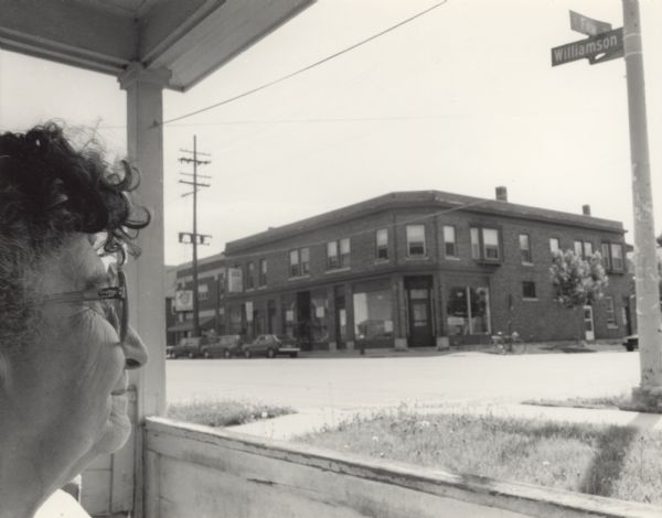 A profile view of the head of a woman, Suzy nee Elvira Dambry, sitting on a porch and looking across the street at a large brick building on the corner of Williamson and Few Street.