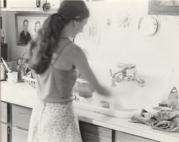 A woman, Karen Barry, is standing over a sink washing dishes. Photographs, paintings, and other small decorations hang on the wall.