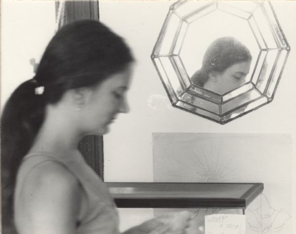 Quarter-length side view portrait of a woman, Karen Barry. Part of her face is reflected in a mirror on the wall. Below the mirror is an aquarium tank with a handwritten sign that reads, in part: "What? A Frog!"