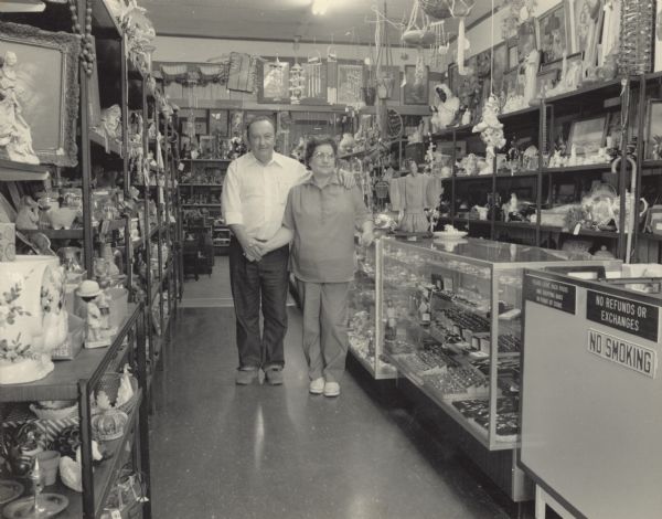 A couple, Doris and Preston Campbell, standing and posing in their store. Preston has his left arm around his wife's shoulders, and the two are holding hands. Doris is resting her right arm on a display case. The room is lined with shelves and cases, holding various objects such as wind chimes, cups, figurines, jewelry, and pictures. Their store is called the "Pantry."