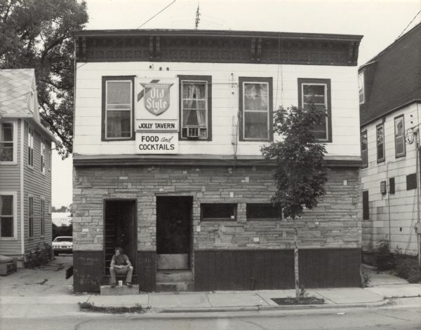 View from across the street of a man, John Cline, sitting on a stoop outside an open door leading to a staircase. There is another doorway on the right. The building displays three signs: "Heileman's Old Style," "Jolly Tavern," and "Food and Cocktails."