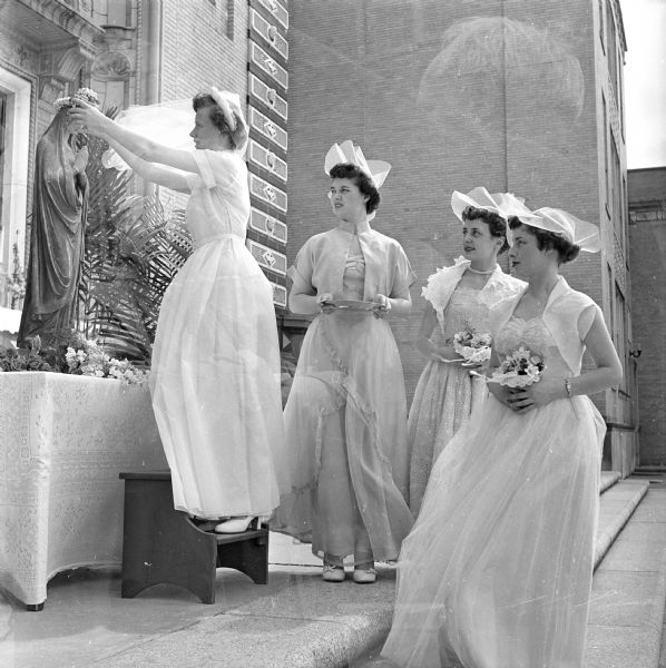 Margaret Dunn placing a floral crown on a statue of the blessed Virgin Mary at the end of the annual May ceremony at Edgewood High School. Other high school seniors in the crowning party are, left to right:  Betty Ferguson, Mary Ann Hand, and Mary Gugel. The double exposure also shows a man in profile against a brick building in the background.