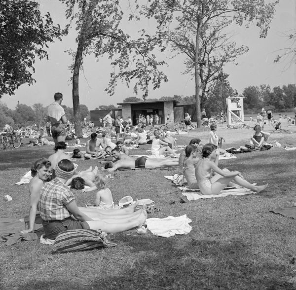 View of a crowd of people, mostly in swimsuits, at what might be Vilas Beach on Lake Wingra. There is a concession stand in a building in the background. A lifeguard sits in a lifeguard chair on the sandy beach on the right.