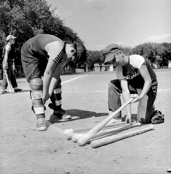 Youth baseball players prepare to play a game. Shown (left to right) are: catcher Edward Allen, Elks Club, who is donning his catcher's equipment, and batter Brad Walrath, Rendall's Clothing, who is selecting his bat.