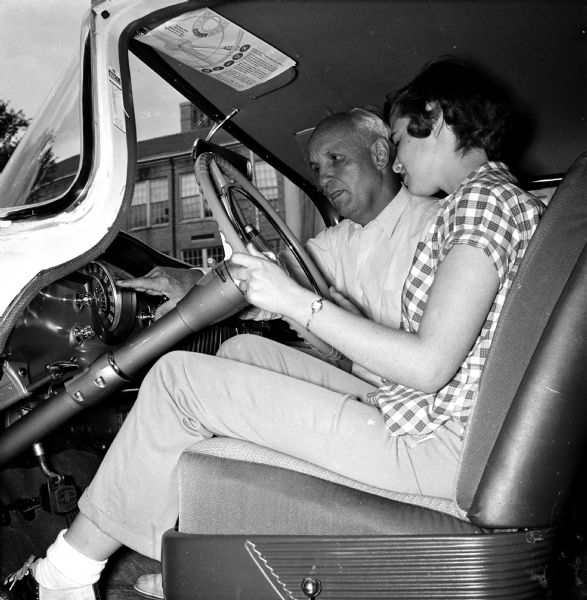 Low angle view of student driver Carol Hansen seated at the steering wheel next to driving instructor and West High School physical education teacher, Willis Jones, who is pointing at the speedometer.