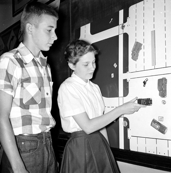Two young people are examining a wall chart depicting traffic lanes, cross walks, and intersections. There are movable adhesive rectangles representing cars. Setting up a traffic problem, is (center) Gail Hawley, while John Pleune (left) is looking on.