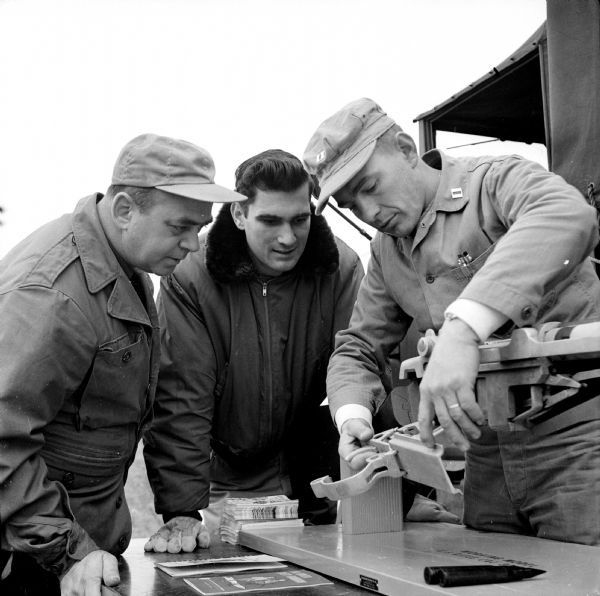 Captain Leo Lewis, inspector-instructor at the Naval-Marine Corps Reserve Training Center in Madison, shows Captain Paul Biehl (left), another instructor, and William Weber of Chicago a wooden model which helps demonstrate the working parts of a rifle. The training session on gun safety was held at the Winnequah Gun Club near Lodi.