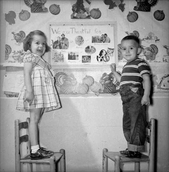 Cynthia Gelman and Sidney Tallard, children in Mrs. V.B. Roberts' nursery school, stand on chairs while inspecting a Thanksgiving poster on the nursery school wall.