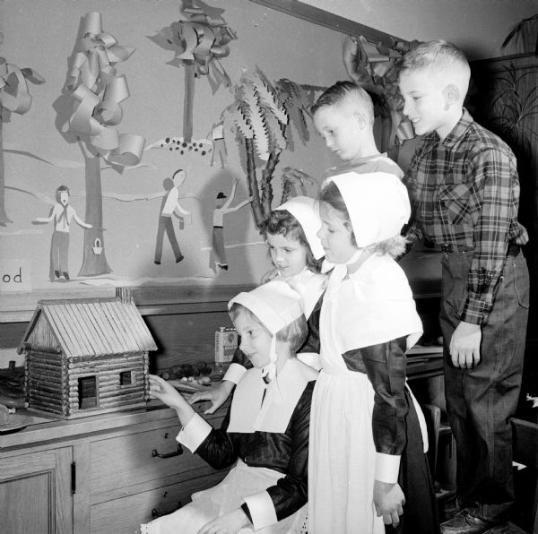 Pupils in Clara Boyle's third grade class at Marquette School wear Puritan costumes while examining a small early American-style log cabin as part of their Thanksgiving study. Seated in front is Beverly Cnare, and behind her are Jillaine Simon, Barbara Sandberg, Michael Kvamme and Michael Banovez.