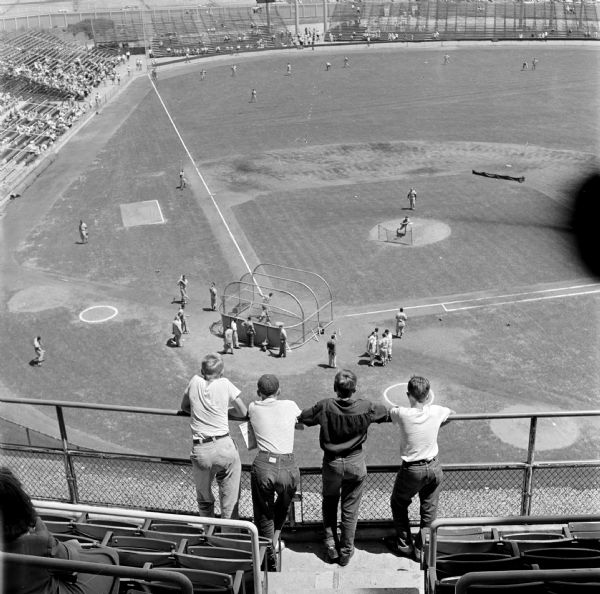 Wisconsin State Journal newspaper carriers attend a Milwaukee Braves versus St. Louis Cardinals baseball game at Milwaukee County Stadium. Four carriers stand along the upper deck railing to get a bird's-eye view of batting practice.