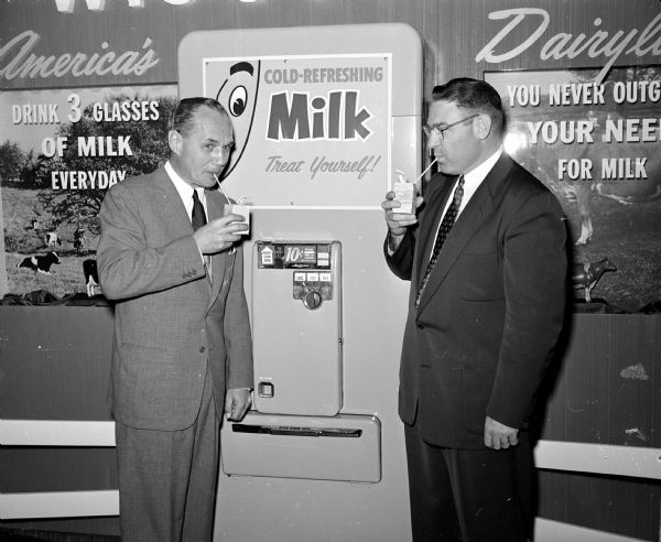 Two men enjoy small cartons of milk with straws in the Capitol building rotunda.  They stand on either side of a tall vending machine with a sign that says, "Cold-Refreshing Milk Treat Yourself Ten Cents." Posters in the background read "From Wisconsin; America's Dairyland; Drink Three Glasses of Milk Everyday; You Never Outgrow Your Need for Milk."