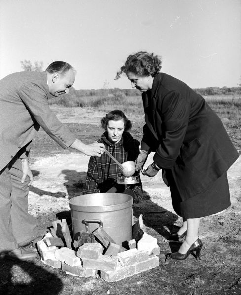 As part of a emergency mass feeding demonstration and course of instruction at Franklin Field, Eldred Heiser ladles soup from a large kettle to Isabelle Hyslop, left, and Janet DePiazza as part of an emergency mass feeding demonstration and course of instruction at Franklin Field. At this two day event, seventy five Madison and Dane County residents will learn what to do if a community's food and water are poisoned by nuclear radiation. The soup pot is sunk into the ground and surrounded by bricks and a fire.