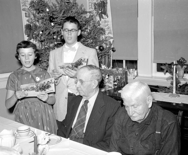 Mary Anderson and Ted Cox, members of the Sherman Avenue Methodist Church youth choir, sing Christmas carols at a party for residents of Oakwood Lutheran Home. Seated at a table are William Clark and August Braemeir.