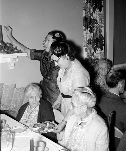 Vivian Schleck (left), member of the Mendota Garden Club, lights a candle, and Mrs. Alvin Sonnenberg,  member of the Little Garden Clubs, serves cookies during a Christmas Party for residents of the Oakwood Lutheran Home. Residents seated at a table are, Nellie Dakins, left and Inger Glimm.