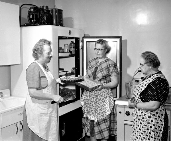 Three ladies are standing in the kitchen while preparing the luncheon for those attending a planning meeting for the upcoming Dane County health survey. Shown are Mrs. Clarence Otto, Sun Prairie; Mrs. August Baumann, Cottage Grove; and Mrs. George T. Mitchell.