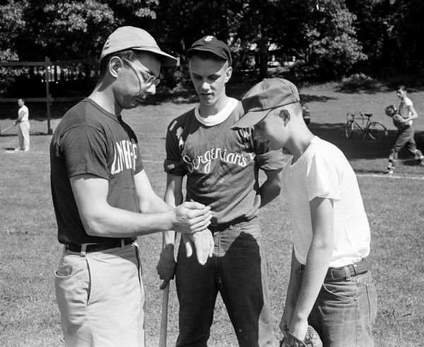 Umpire Mel Gaestel flips a coin to start the baseball game. Players George Oosterhaus from the Sergenian's Carpet team and Dan Olson from the Optimists team look on.