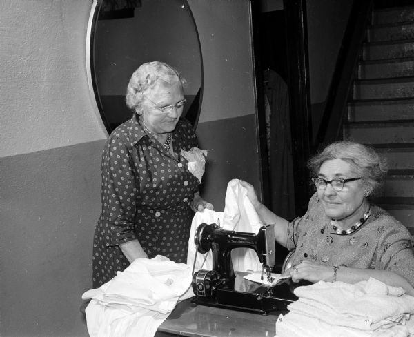 Two women, one standing, and the other sitting by a sewing machine "are examining some of the men's shirts which have been converted into bed jackets for a Madison Home for the Aged."  The women are Mrs. W.F. Adolphsen (standing) and Ella Luick. The original caption states: "members of the Sewing circle of the Luther Memorial church gather at the church frequently to sew for various needy groups."