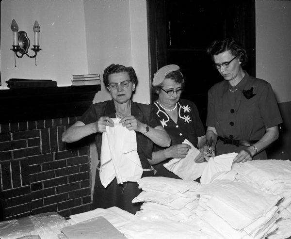 Three women prepare "materials used in the making of cancer dressings."  The women include Eleanor Britt, Florence Anderson, and Marcia Udelhofen. The original caption states, "members of the Sewing circle of the Luther Memorial church gather at the church frequently to sew for various needy groups."
