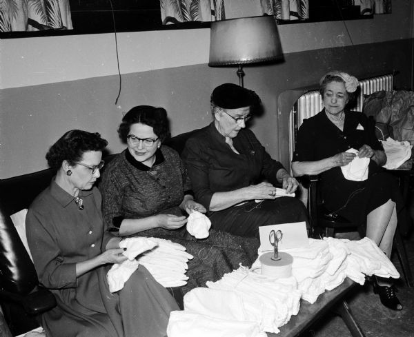 Four women are sitting together behind a low table containing small bundles of cloth and a scissors while making slippers for the children staying at a Madison hospital. The women are Carolyn Breitzke, Emma Hooper, Caroline Schwingel, and Lillian Hoernke.  The original caption states: "members of the Sewing circle of the Luther Memorial church gather at the church frequently to sew for various needy groups."