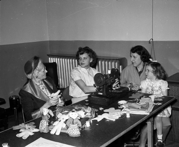 Three women and a girl are sitting and chatting at a table while making toys for a Madison hospital. Shown as they sew and stuff cloth toys, are (left to right:) Hattie Bakke, Doris Marten,and Helen Schaeve, who is holding her daughter, Carolyn. The original caption states: "members of the Sewing circle of the Luther Memorial Church gather at the church frequently to sew for various needy groups."
