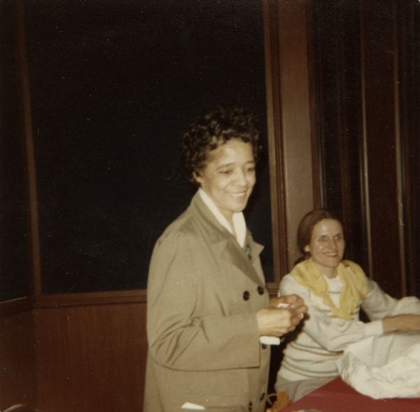Vel Phillips, wearing a tan coat, is standing at a table, with another woman sitting behind her.