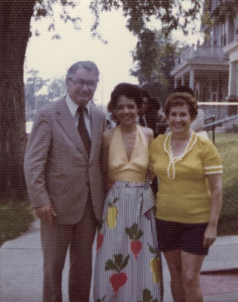 Vel Phillips posing outdoors on a sidewalk with Patrick Lucey and a woman on the right. A group of people stand behind them. Vel is wearing a yellow top over a long, striped skirt with a vegetable design. In the background is a house on the right and a tree on the left.