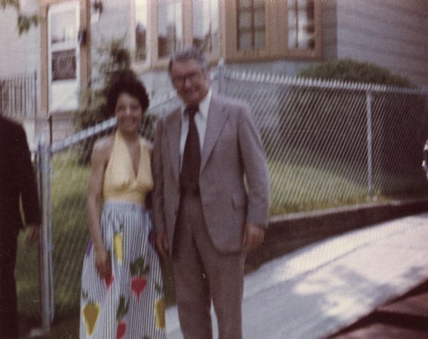 Vel Phillips posing outdoors with a man wearing a suit. Vel is wearing a yellow top over a long, striped skirt with a vegetable design. Behind them is a driveway, a fence, and a house.