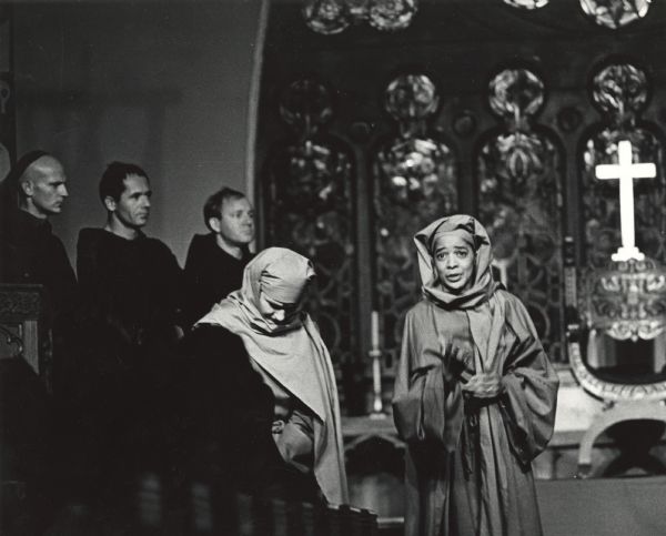 Vel Phillips standing and speaking in a costume. There is a woman on the left bowing her head. Three men are standing on the left looking to the right. In the background is a large cross and stained glass windows. This image is from the 1972 Milwaukee production of T.S. Eliot's play "Murder in the Cathedral."