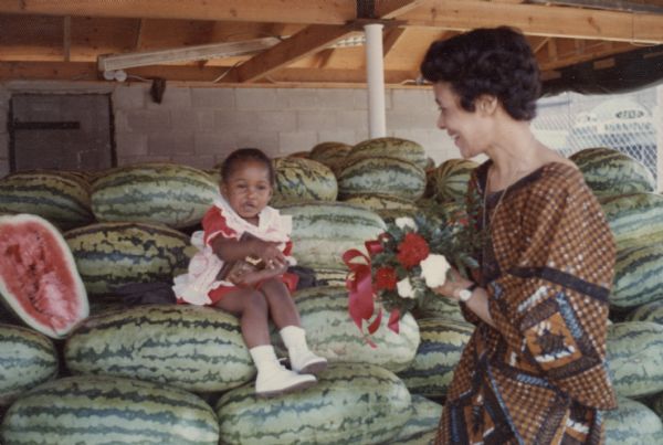 Vel Phillips, wearing a brown and blue patterned dress is holding a bouquet of flowers. She stands near a young girl wearing a dress sitting on a pile of watermelons and holding a plaque in her lap.