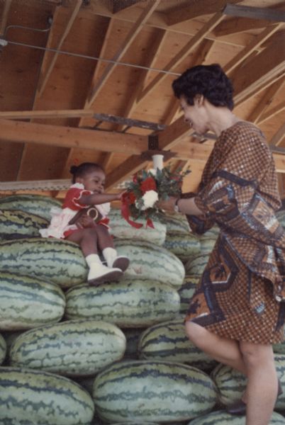 Vel Phillips, wearing a brown and blue patterned dress is standing and holding a bouquet of flowers. A young girl sitting on a pile of watermelons holds a plaque in her lap while reaching out towards the flowers.