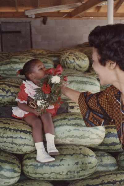 Vel Phillips is in a brown and blue patterned dress standing and holding a bouquet of flowers up to a young girls' nose. The girl is holding a plaque in her lap and is sitting on a pile of watermelons.