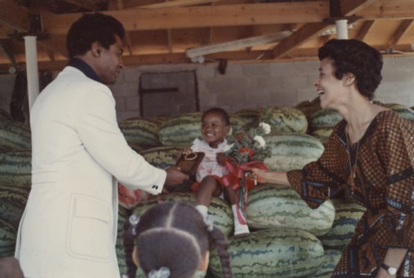 Vel Phillips, wearing a brown and blue patterned dress, is standing on the right holding a bouquet of flowers. A unidentified man is standing on the left, holding on to a plaque that a young girl is also holding. The girl is sitting on a pile of watermelons. There is another child in the foreground with hair ties in her hair.