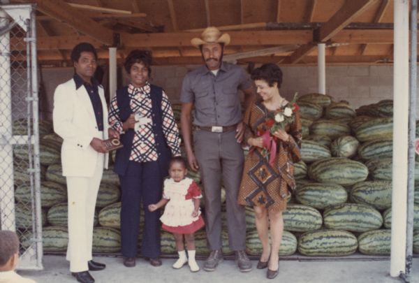 Vel Phillips, standing on right, is in a brown and blue patterned buba (a Yoruba blouse) and holding a bouquet of flowers. Next to her (second from right) is an unidentified man in blue work clothes and a hat. A young girl stands in the center wearing a red and white dress. An unidentified woman (second from left) is wearing a blue and red striped long sleeve shirt with a blue vest. An unidentified man (left) is in a white suit and is holding a plaque. There is another child in the bottom left corner facing the group. In the background is a stack of watermelons in an enclosure.