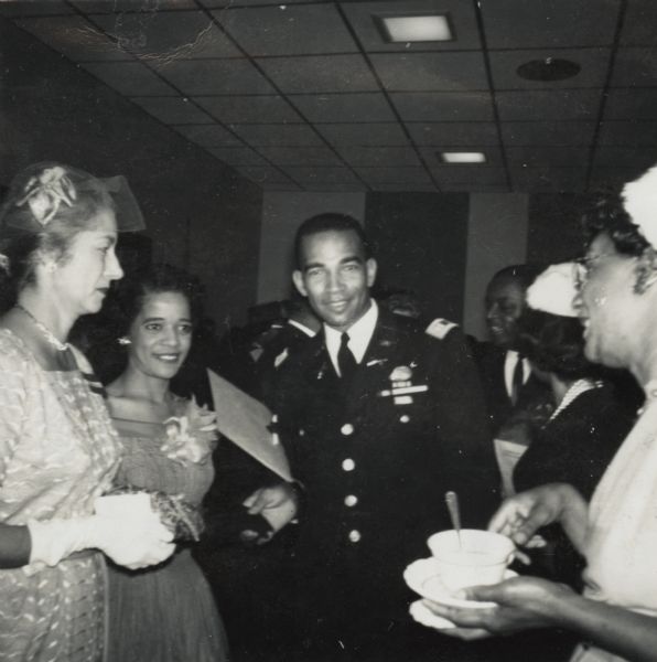 Vel Phillips (second to left) is standing and wearing a dress with a flower brooch. Next to her is Captain Al Roper in military uniform. Two unidentified women stand near them in the foreground. Other unidentified people are in the background.