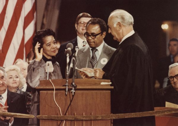 Vel Phillips, wearing a grey outfit is standing at a podium holding up her right hand near Wisconsin Supreme Court Chief Justice Bruce Beilfuss standing on the right. In between them is Vel's husband W. Dale Phillips. In the bottom left corner is Wisconsin Governor Lee S. Dreyfus.